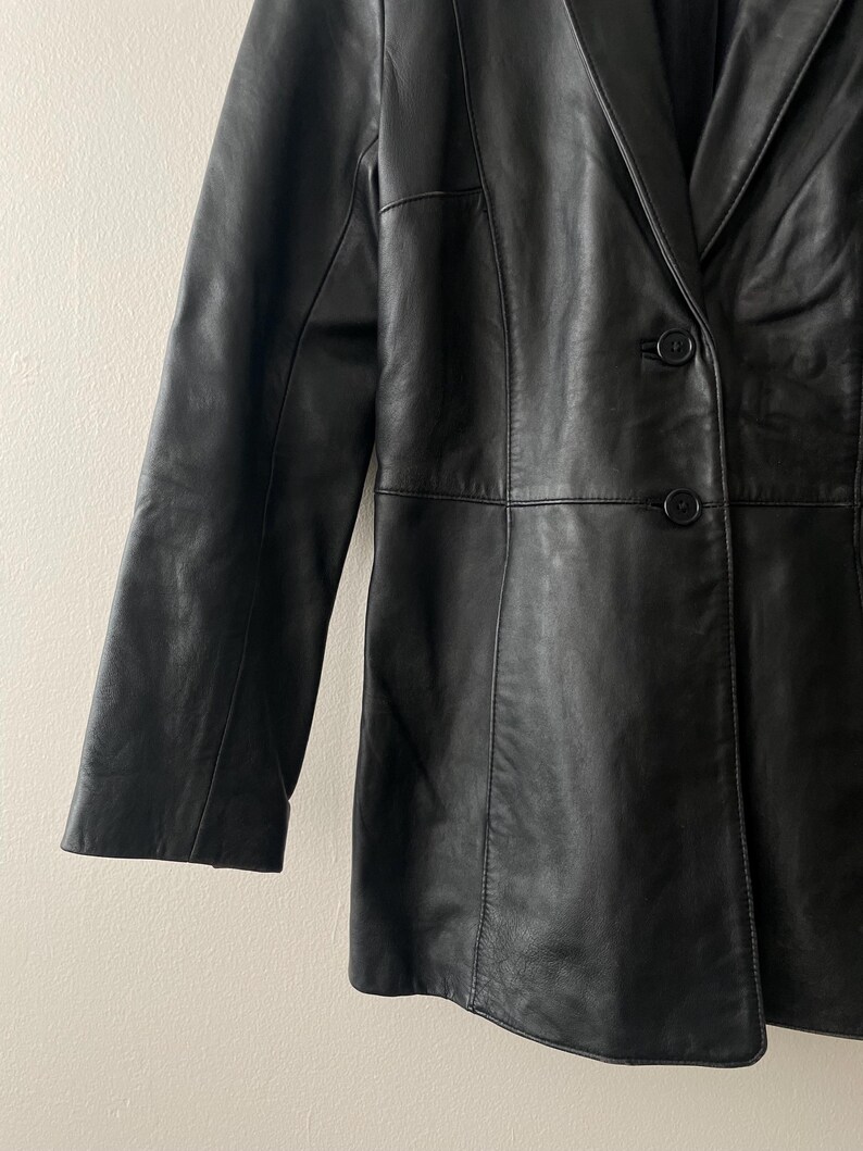 May include: A black leather blazer with a classic design. The blazer features a notched lapel, two-button closure, and long sleeves. The leather appears to be smooth and has a slight sheen. The garment is tailored with seams for a structured fit.