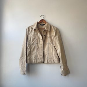 May include: Tan corduroy jacket with button closures, a collared neckline, and front pockets. The jacket is hanging on a wooden hanger against a neutral background. Features visible stitching details.