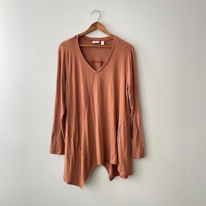 May include: Rust-colored long-sleeve shirt with a V-neck and asymmetrical hem. The shirt features horizontal stripes in a lighter shade of orange. The garment is hanging on a wooden hanger against a plain white wall.