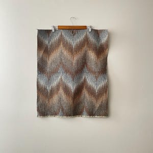 May include: A rectangular textile with a brown, gray, and blue chevron pattern hangs from a wooden hanger. The fabric has a fringed edge at the bottom. The design is a repeating zig-zag pattern.