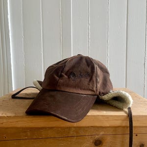 May include: A brown baseball cap with a curved peak and ear flaps lined with cream-coloured faux shearling. The front of the cap features the word Outback embroidered in dark brown thread. Long brown straps extend from the sides of the ear flaps.