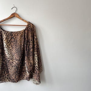 Könnte beinhalten: Ein Oberteil mit Leopardenmuster und langen, fließenden Ärmeln hängt an einem Holzbügel. Das Oberteil hat einen Rundhalsausschnitt und ein braunes, beiges und schwarzes Fleckenmuster. Das Kleidungsstück ist aus leichtem Material gefertigt.