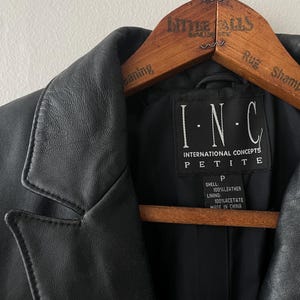 May include: Black leather jacket with a notched collar, hanging on a wooden hanger. The label reads "I.N.C. INTERNATIONAL CONCEPTS PETITE". The jacket is made of 100% leather and 100% acetate lining.