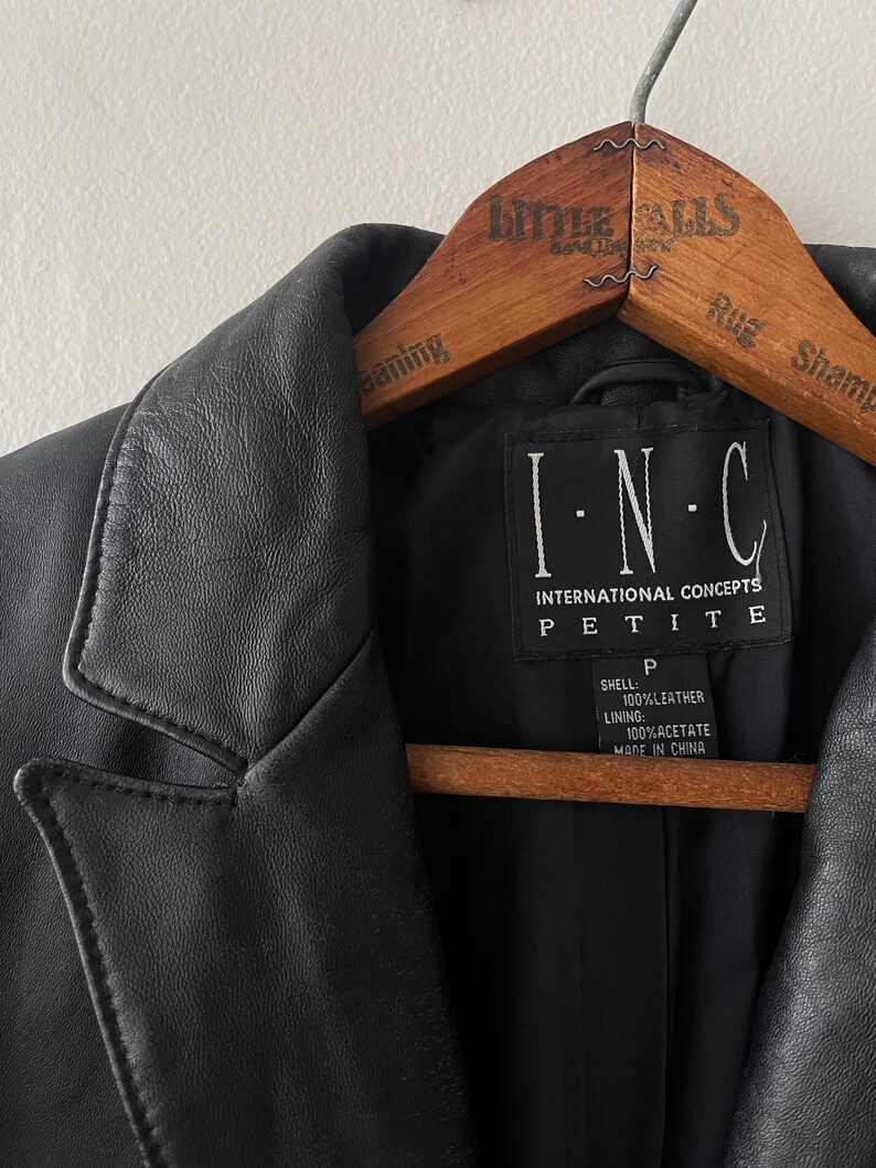 May include: Black leather jacket with a notched collar, hanging on a wooden hanger. The label reads "I.N.C. INTERNATIONAL CONCEPTS PETITE". The jacket is made of 100% leather and 100% acetate lining.