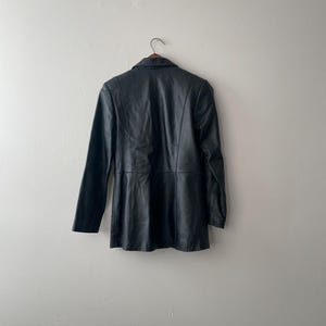 May include: A black leather blazer hanging on a wooden hanger. The blazer has a classic collar, long sleeves, and a tailored fit. The garment is displayed against a plain, off-white wall.