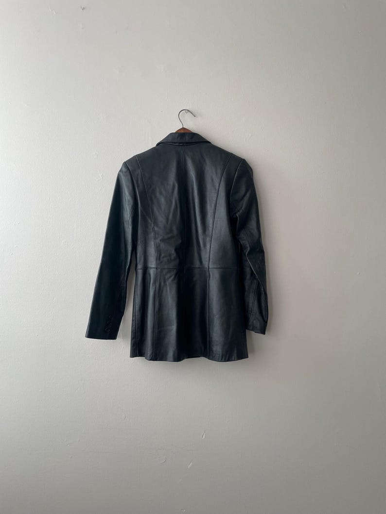 May include: A black leather blazer hanging on a wooden hanger. The blazer has a classic collar, long sleeves, and a tailored fit. The garment is displayed against a plain, off-white wall.