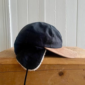 May include: A black cap featuring a tan suede brim and fold-down ear flaps lined with white faux shearling. The cap has a rounded crown and a thin black strap hanging from the side, providing coverage for the ears and head in cold weather.