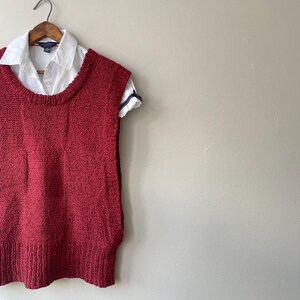 May include: A red knitted vest with a crew neck is displayed on a wooden hanger. A white collared shirt is layered underneath, with the sleeves visible. The shirt has a small logo on the chest. The vest has a textured appearance.