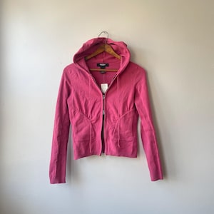May include: A bright pink hooded zip-up sweater with long sleeves. The sweater has a fitted silhouette with a drawstring detail at the waist and two front pockets. The sweater is hanging on a wooden hanger.