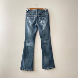 May include: Faded blue denim jeans with a flared leg design. The jeans have decorative stitching on the back pockets and a classic five-pocket design. The jeans are hanging on a wooden hanger.