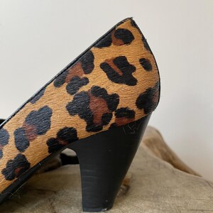 May include: A high-heeled shoe featuring a textured leopard print pattern in shades of tan, brown, and black. The shoe has a solid black heel and black trim along the upper edge, showcasing a distinct animal-inspired design on the exterior.