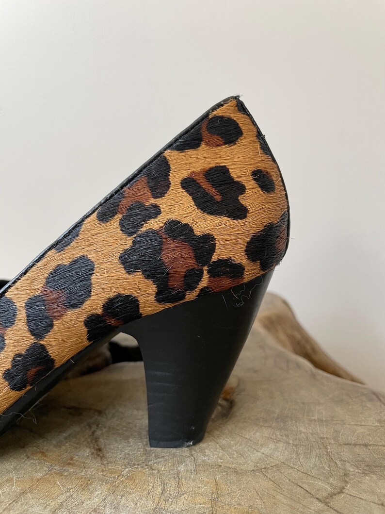 May include: A high-heeled shoe featuring a textured leopard print pattern in shades of tan, brown, and black. The shoe has a solid black heel and black trim along the upper edge, showcasing a distinct animal-inspired design on the exterior.
