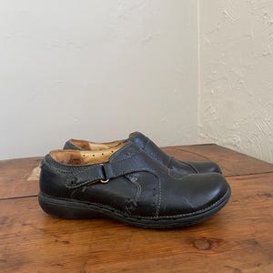 May include: A pair of black leather slip-on shoes with a buckle closure. The shoes have visible stitching and a rounded toe. The soles are dark and the shoes appear worn. The shoes are on a wooden surface.