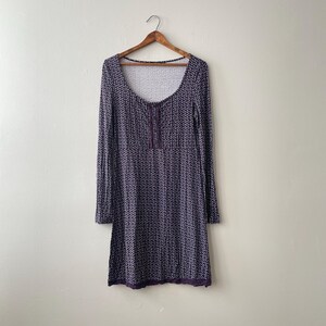 Pode incluir: Vestido de manga comprida com decote redondo e padrão geométrico roxo escuro e branco. O vestido tem um detalhe de botões no peito e uma barra de renda roxa escura na bainha. Pendurado em um cabide de madeira.