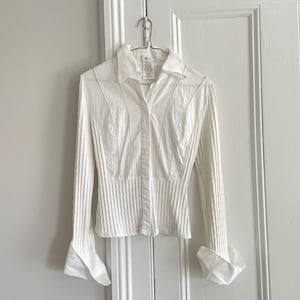 May include: A white long-sleeve button-down shirt featuring a structured collar, vertical ribbed fabric panels on the sides and sleeves, smooth fabric panels on the front, and wide, flared cuffs at the wrists, all hanging on a metal wire hanger.
