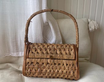 May include: A rectangular woven basket handbag with a curved handle. The bag is made of light brown woven material with a button closure. The handle is also made of woven material.