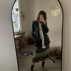 May include: A full-length mirror with a black frame reflects a person wearing a black leather jacket over a white shirt, a floral print skirt, black socks, and tan slip-on shoes. The mirror is arched at the top.