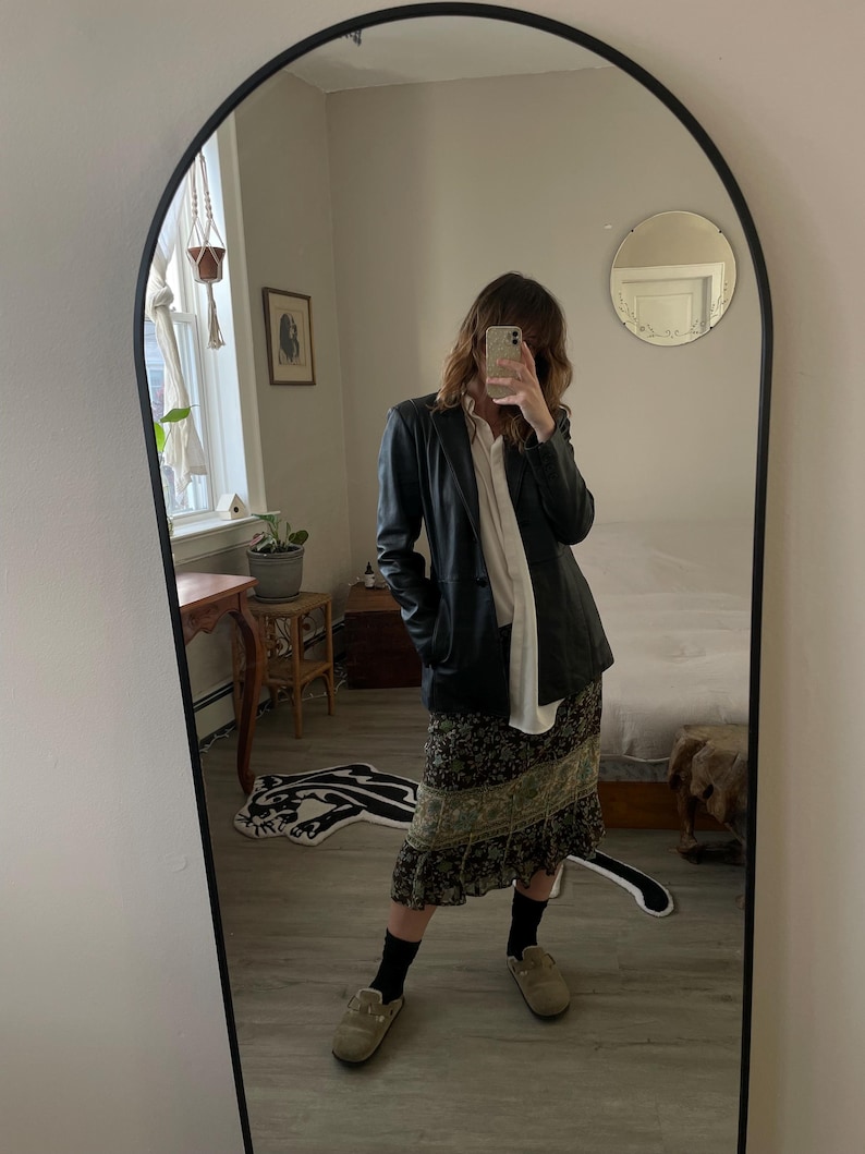 May include: A full-length mirror with a black frame reflects a person wearing a black leather jacket over a white shirt, a floral print skirt, black socks, and tan slip-on shoes. The mirror is arched at the top.