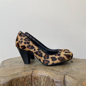 May include: A pair of high-heeled pumps featuring a textured leopard print pattern in shades of brown, tan, and black. The shoes have a rounded toe, a black interior lining, and a solid black block heel that provides a sturdy base for the footwear.