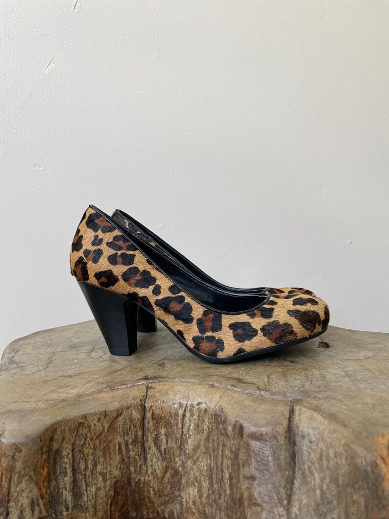 May include: A pair of high-heeled pumps featuring a textured leopard print pattern in shades of brown, tan, and black. The shoes have a rounded toe, a black interior lining, and a solid black block heel that provides a sturdy base for the footwear.