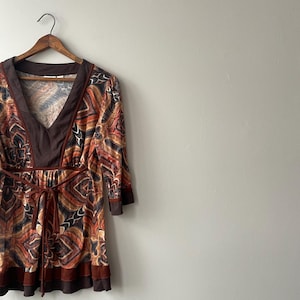 May include: A patterned tunic top in shades of brown, orange, and black hangs on a wooden hanger. The top features a V-neck, three-quarter sleeves, and a tie at the waist. The fabric has a geometric design.