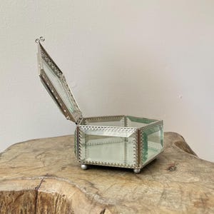 May include: A hexagonal glass jewelry box with a hinged lid and silver-toned metal trim. The box features clear glass panels, a mirrored base, and small spherical feet. The metal edges are decorated with a delicate, repeating scalloped pattern.