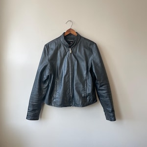 May include: Black leather jacket with a zip-up front and a stand-up collar. The jacket is hanging on a wooden hanger against a neutral wall. The jacket has a classic, streamlined design.