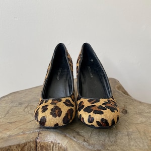 May include: A pair of BCBGMAXAZRIA pumps featuring a textured leopard print pattern in shades of tan, brown, and black. These shoes have a rounded toe, a black interior lining, and a high heel, showcasing a classic silhouette with a bold animal print.