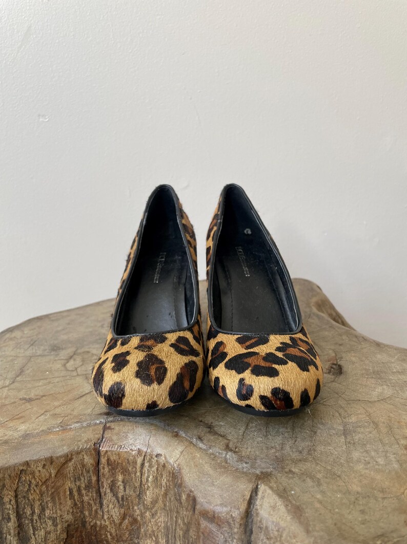 May include: A pair of BCBGMAXAZRIA pumps featuring a textured leopard print pattern in shades of tan, brown, and black. These shoes have a rounded toe, a black interior lining, and a high heel, showcasing a classic silhouette with a bold animal print.