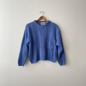 May include: A periwinkle blue long-sleeve sweater featuring a chunky cable knit pattern, a crew neckline, and a single patch pocket on the left chest. The interior collar tag displays the brand name Forenza in black text on a white background.