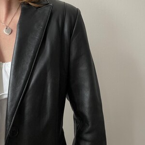 May include: Black leather blazer with a single-button closure. The blazer has a classic lapel and a tailored fit. The image shows the blazer's shoulder and sleeve detail, showcasing the smooth texture of the leather. A silver heart pendant necklace is visible.