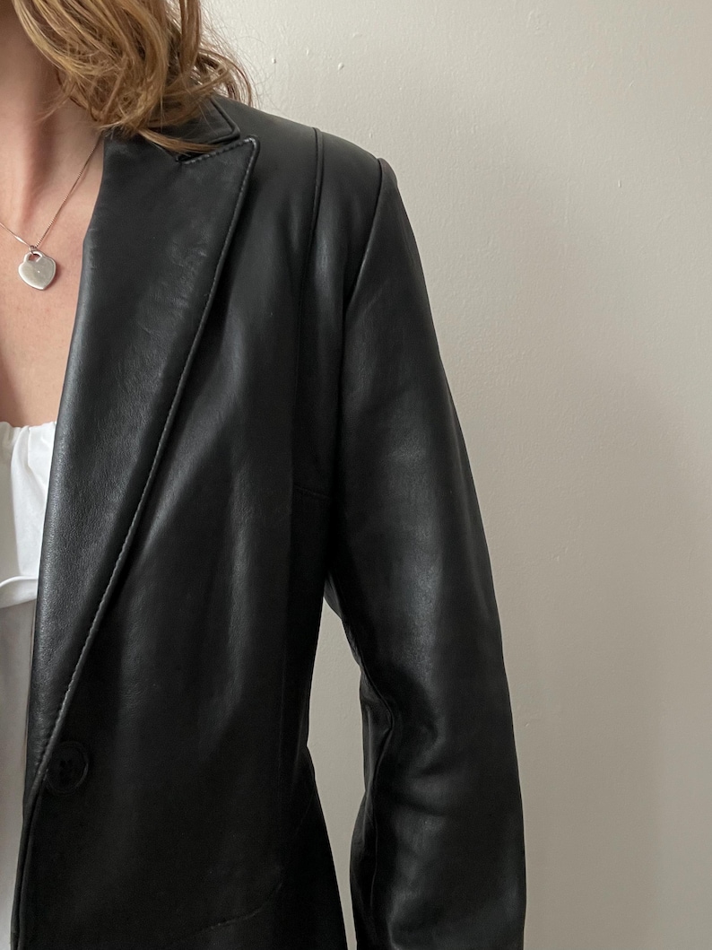 May include: Black leather blazer with a single-button closure. The blazer has a classic lapel and a tailored fit. The image shows the blazer's shoulder and sleeve detail, showcasing the smooth texture of the leather. A silver heart pendant necklace is visible.