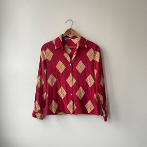 May include: A red button-down shirt with a repeating pattern of tan diamond shapes. The long-sleeved shirt is hanging on a wooden hanger against a plain white background. The shirt has a collared neckline and button closures.