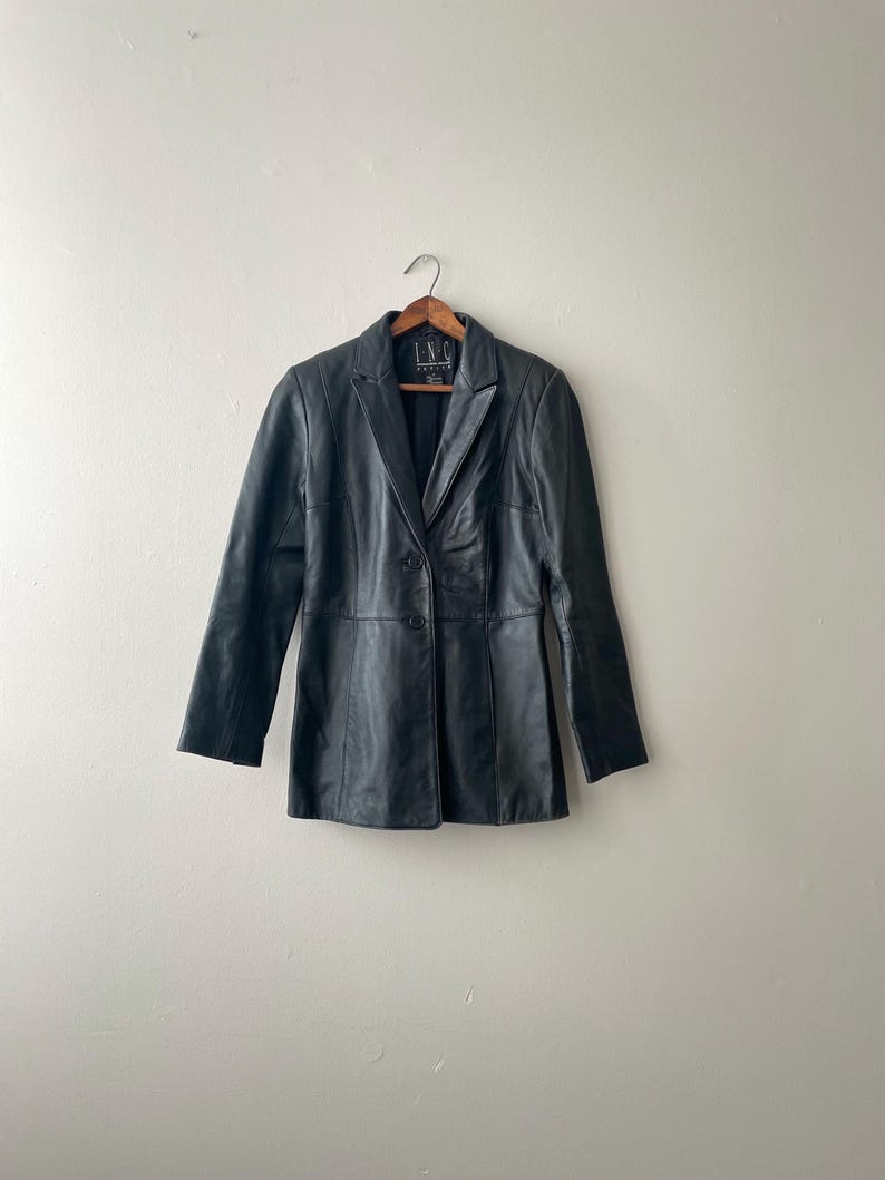 May include: A black leather blazer with a notched collar and two-button closure. The blazer is hanging on a wooden hanger against a neutral background. The blazer has a tailored fit and long sleeves.