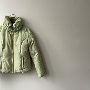 Puede incluir: Chaqueta acolchada verde claro con cuello alto fruncido. La chaqueta tiene cierre de cremallera y dos bolsillos delanteros. La tela tiene un patrón sutil y texturizado. La chaqueta está colgada de una percha de madera.