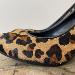 May include: A high-heeled shoe featuring a textured leopard print pattern in shades of tan, brown, and black. The shoe has a rounded toe, a black interior lining, and a black trim along the opening, showcasing a detailed animal-inspired design.