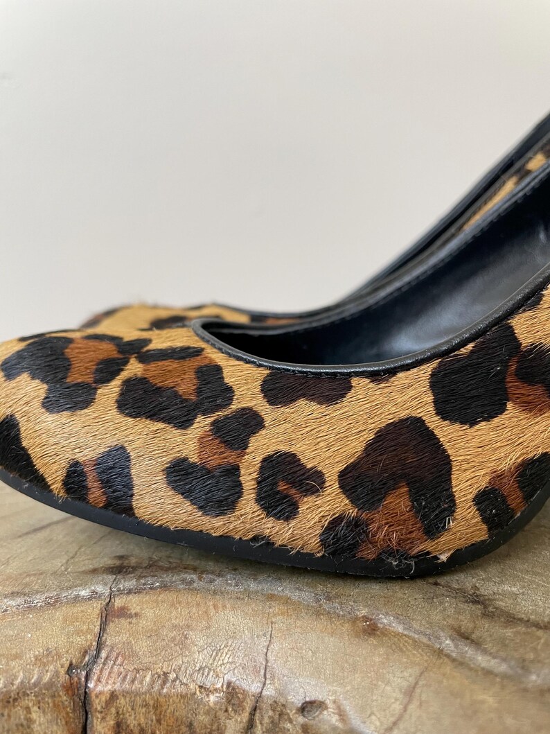 May include: A high-heeled shoe featuring a textured leopard print pattern in shades of tan, brown, and black. The shoe has a rounded toe, a black interior lining, and a black trim along the opening, showcasing a detailed animal-inspired design.