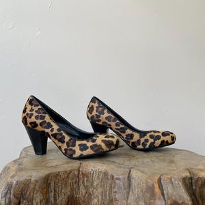 May include: A pair of pumps featuring a textured leopard print pattern in shades of tan, brown, and black. The shoes have a rounded toe, a black interior lining, and a solid black block heel. The exterior material has a short, fur-like texture.