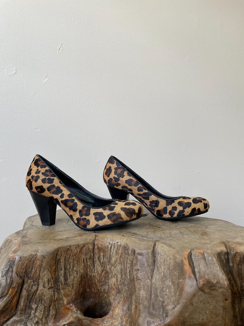 May include: A pair of pumps featuring a textured leopard print pattern in shades of tan, brown, and black. The shoes have a rounded toe, a black interior lining, and a solid black block heel. The exterior material has a short, fur-like texture.