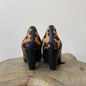 May include: A pair of high-heeled shoes featuring a textured leopard print pattern in shades of brown and black. The shoes have a solid black vertical seam running down the back of the heel and a smooth black heel base for stability and support.