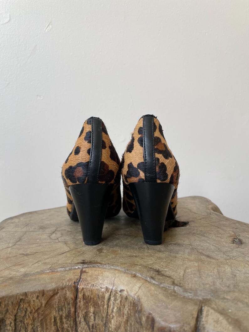 May include: A pair of high-heeled shoes featuring a textured leopard print pattern in shades of brown and black. The shoes have a solid black vertical seam running down the back of the heel and a smooth black heel base for stability and support.