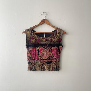 May include: A sleeveless crop top featuring a black base with a vibrant, multi-colored paisley and floral pattern in shades of hot pink, gold, and yellow. The garment has a wide, rounded neckline and a straight hem, hanging on a wooden hanger.