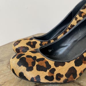May include: A pair of high-heeled pumps featuring a textured leopard print pattern in shades of tan, brown, and black. The shoes have a rounded toe, a black interior lining, and a black trim along the opening of the shoe upper.