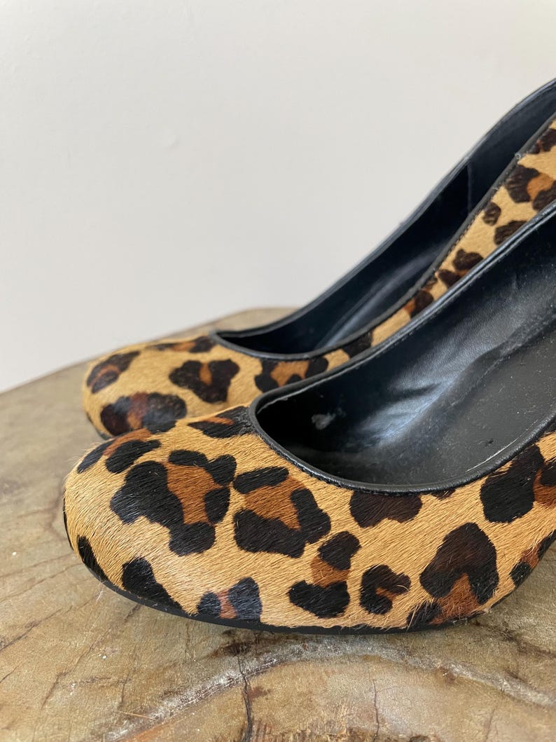 May include: A pair of high-heeled pumps featuring a textured leopard print pattern in shades of tan, brown, and black. The shoes have a rounded toe, a black interior lining, and a black trim along the opening of the shoe upper.