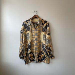 May include: A button-down shirt with a gold, black, and gray pattern. The long-sleeved shirt features a repeating design of ornate shapes and crests. The shirt is hanging on a wooden hanger against a plain wall.