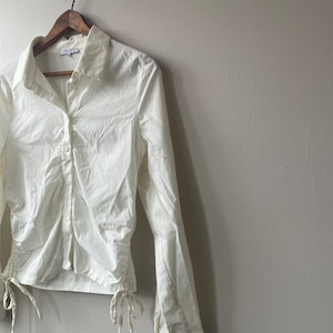 Puede incluir: Una camisa blanca de manga larga con cuello. La camisa tiene una silueta entallada con detalles de cordón en la cintura y los puños. Cuelga de una percha de madera contra una pared neutra.
