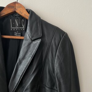 May include: Black leather blazer hanging on a wooden hanger. The blazer has a notched lapel, a single-button closure, and a petite fit. The label reads "I.N.C. INTERNATIONAL CONCEPTS PETITE".