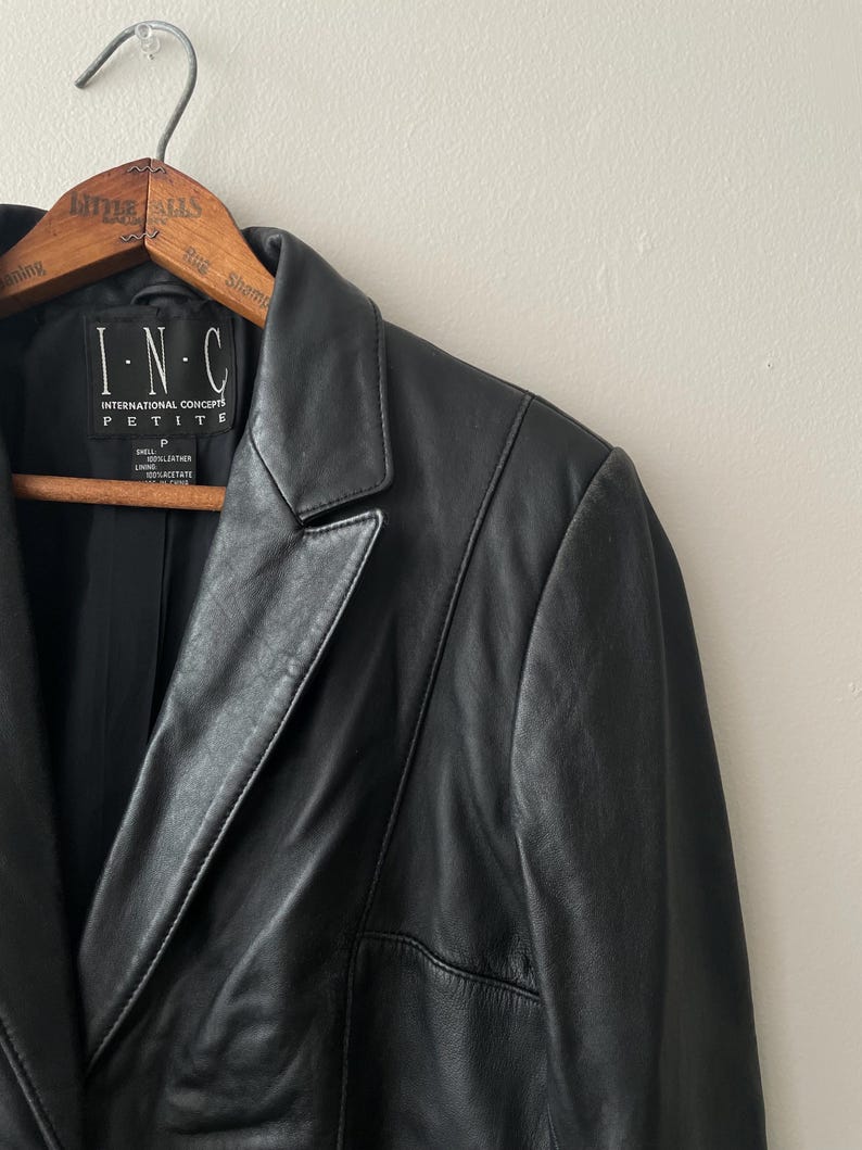 May include: Black leather blazer hanging on a wooden hanger. The blazer has a notched lapel, a single-button closure, and a petite fit. The label reads "I.N.C. INTERNATIONAL CONCEPTS PETITE".