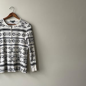 May include: White and navy blue long-sleeve Henley shirt with a geometric pattern. The shirt is hanging on a wooden hanger. The shirt has a button-up neckline and a ribbed cuff.