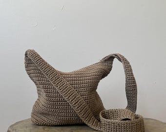 Könnte beinhalten: Eine braune gehäkelte Umhängetasche mit passendem Armband. Die Tasche hat einen breiten Riemen und ein einzigartiges, strukturiertes Design. Tasche und Armband sind aus weichem, gewebtem Material und liegen auf einer Holzoberfläche.